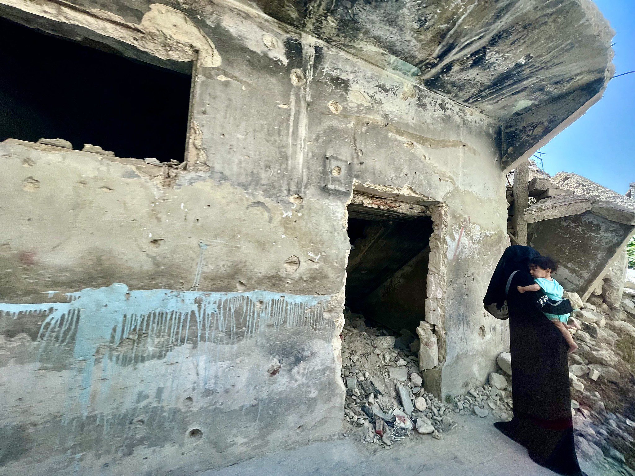 Photo of a woman dressed in black holding baby in front of a concrete building and rubble.