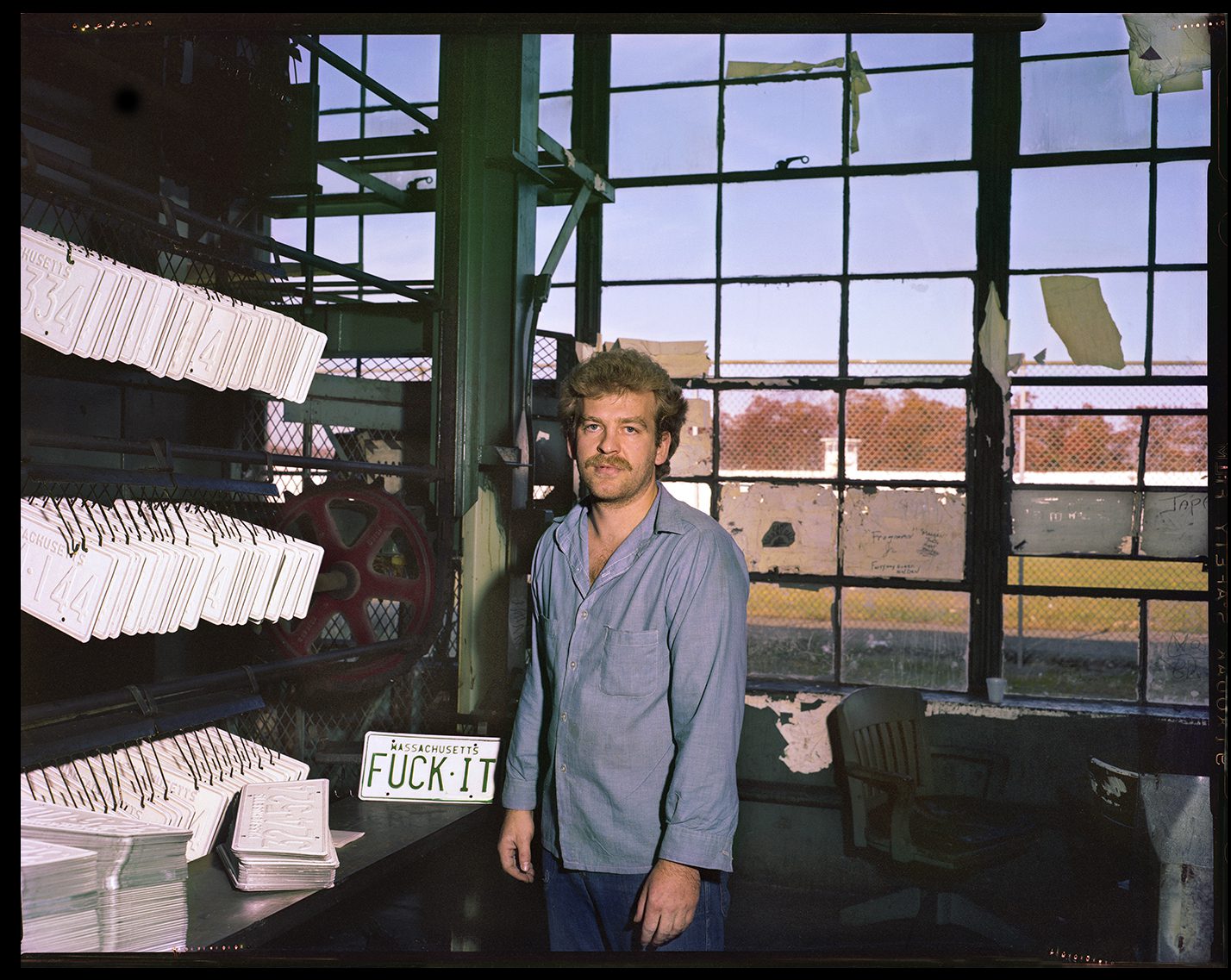 A man stands before a large window, and machinery to make license plates. One plate reads, "FUCK-IT".
