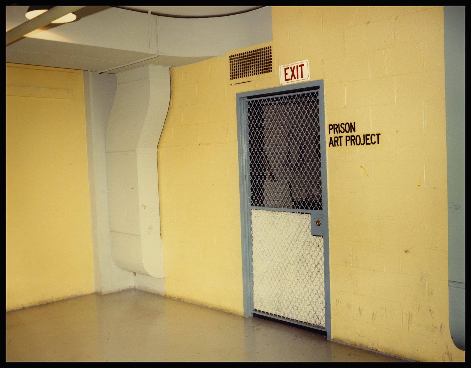 A caged door with an exit sign over it, the words "Prison Art Project" painted on the yellow wall.