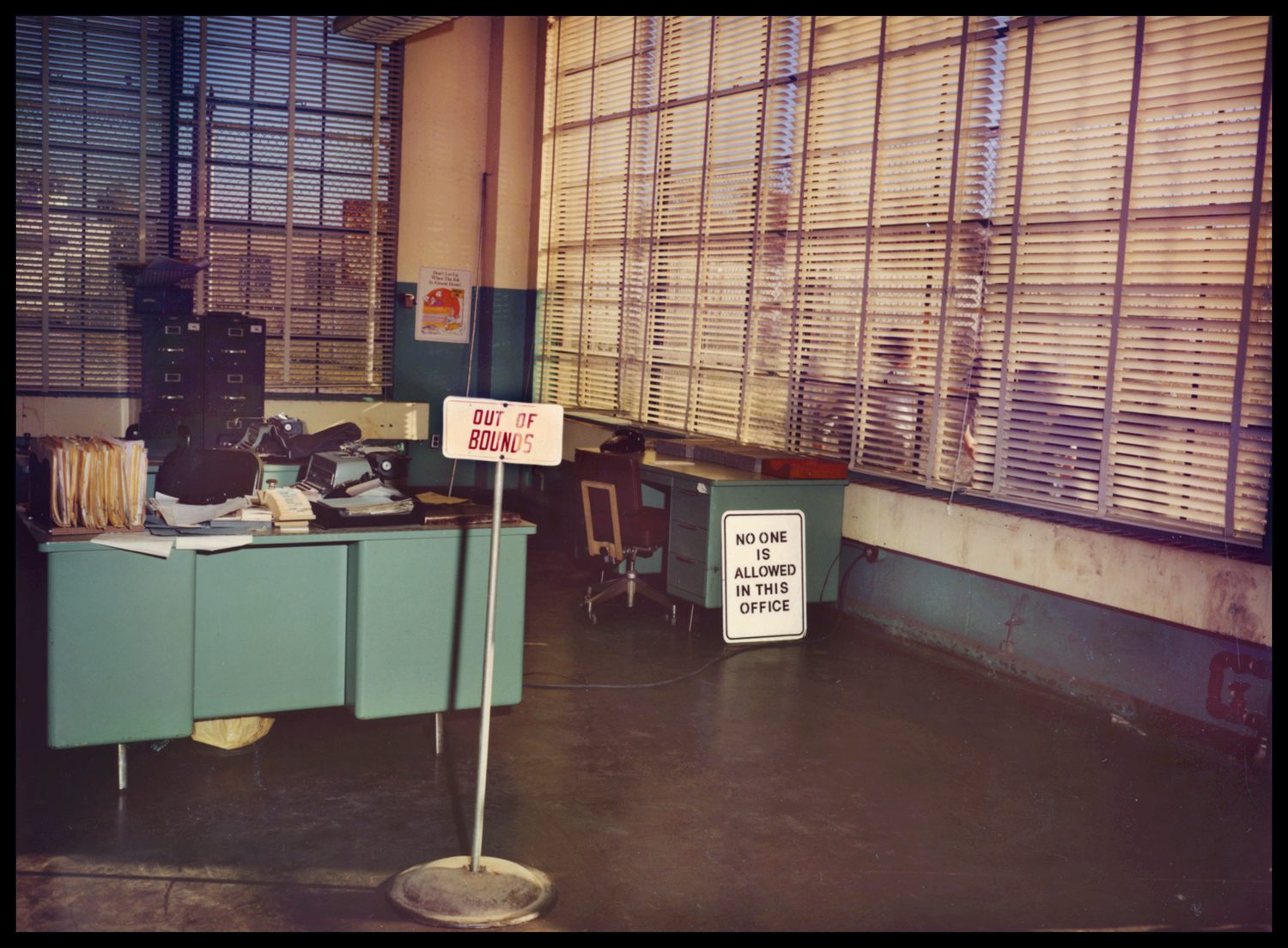 2 desks by large windows with blinds, signs read "Out of bounds" and "no one is allowed in this office."