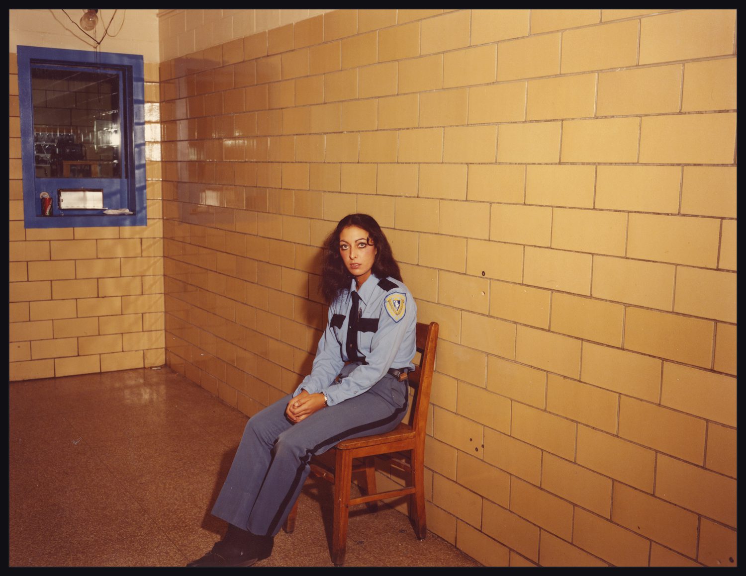 A woman in a guard uniform sits in a chair, against a faded yellow tile wall