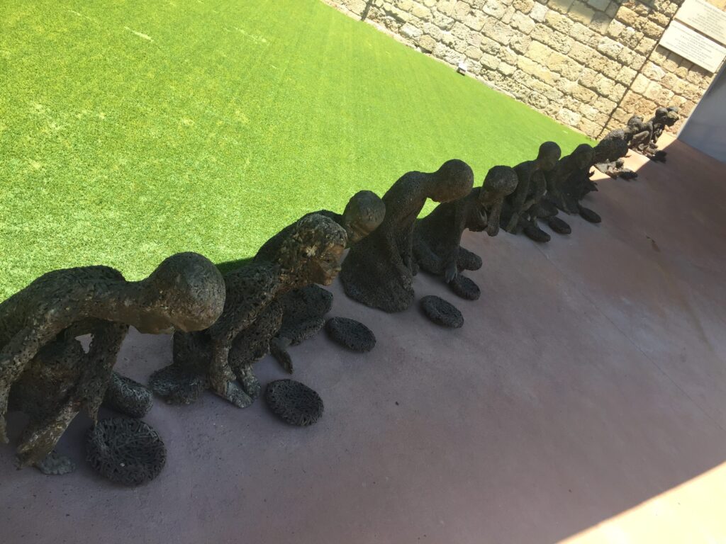 Arc of green grass in the the sun, line of statues of small children with empty bowls in the shade.