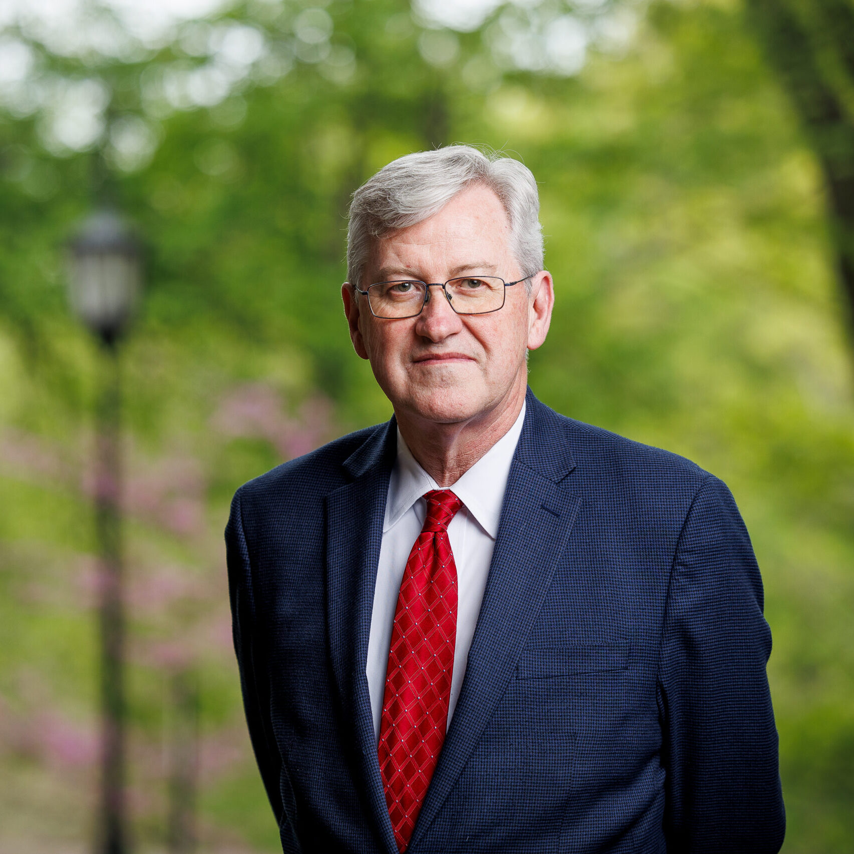 photo of white-haired man wearing blue suit and red tie against out of focus trees