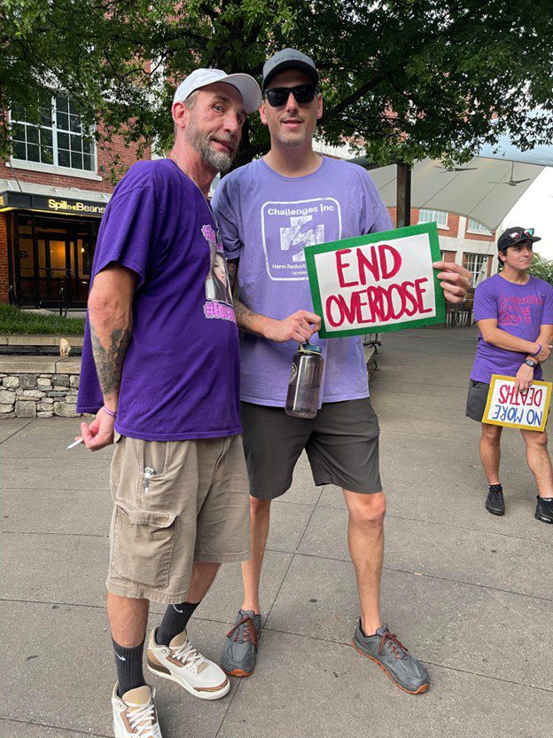 Photo of two men in purple tshirts and shorts. One holds a sign that says "end overdose"