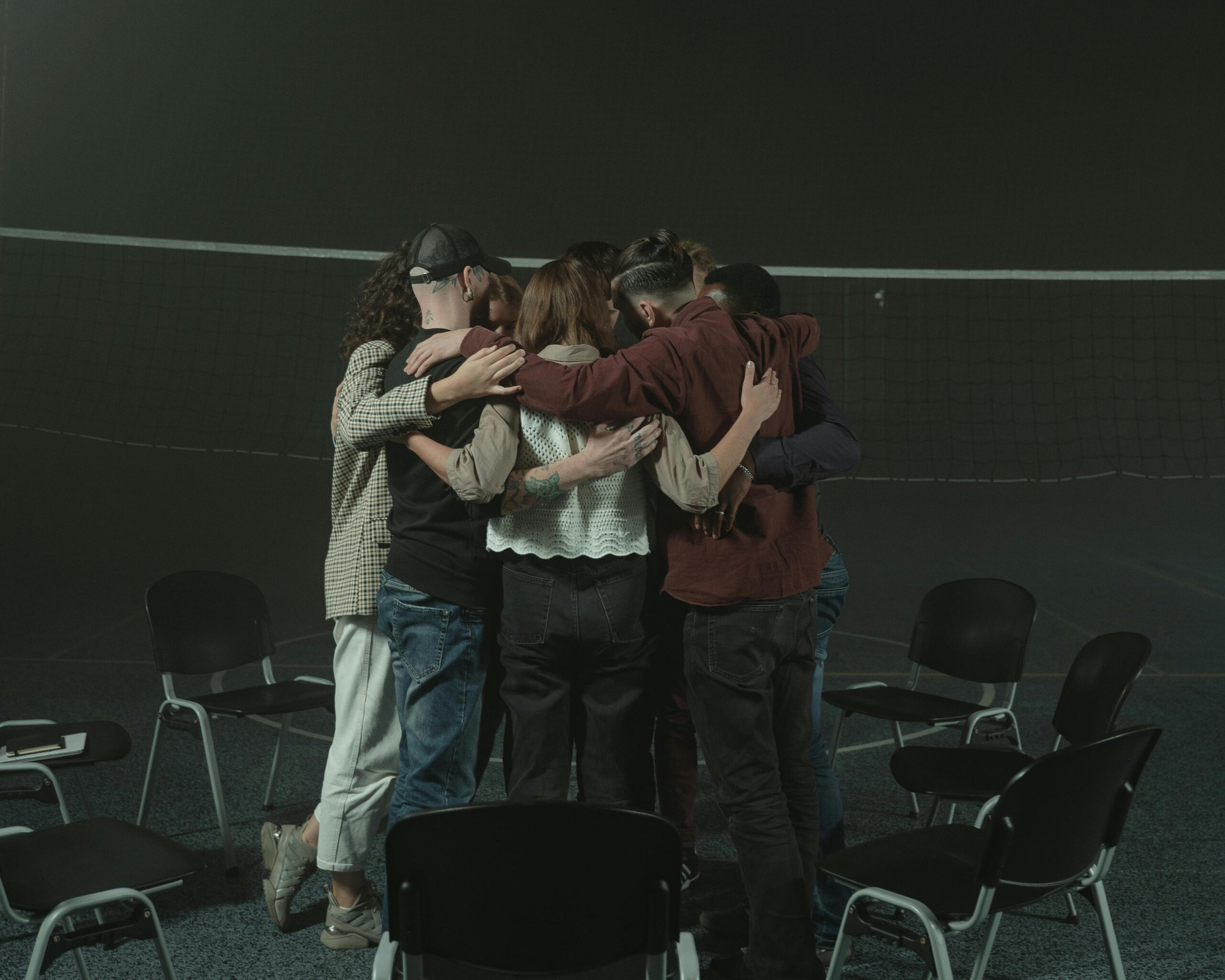 Photo back view of a group of individuals hugging in a circle surrounded by chairs.