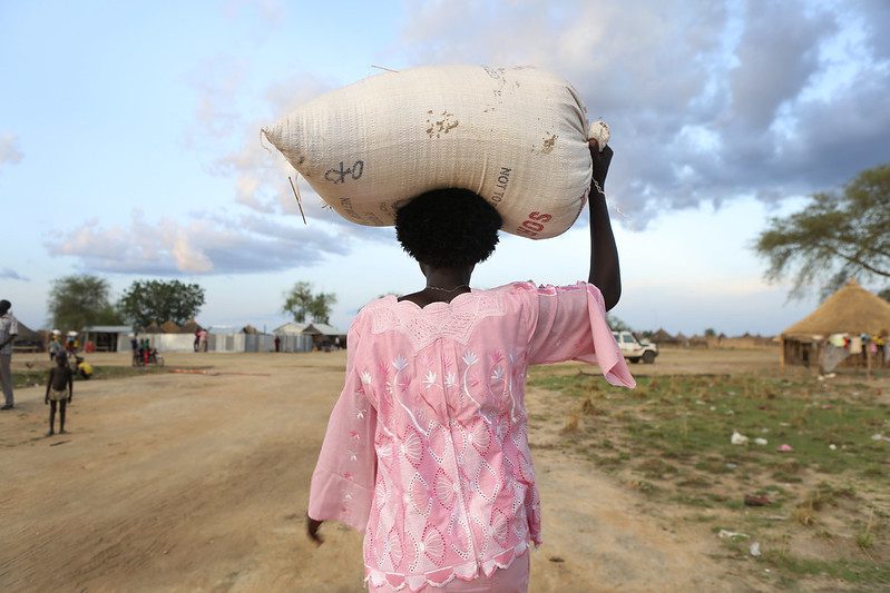 photo back view of a woman in a pink shirt carrying a sack of food on her head walking down a dirt road