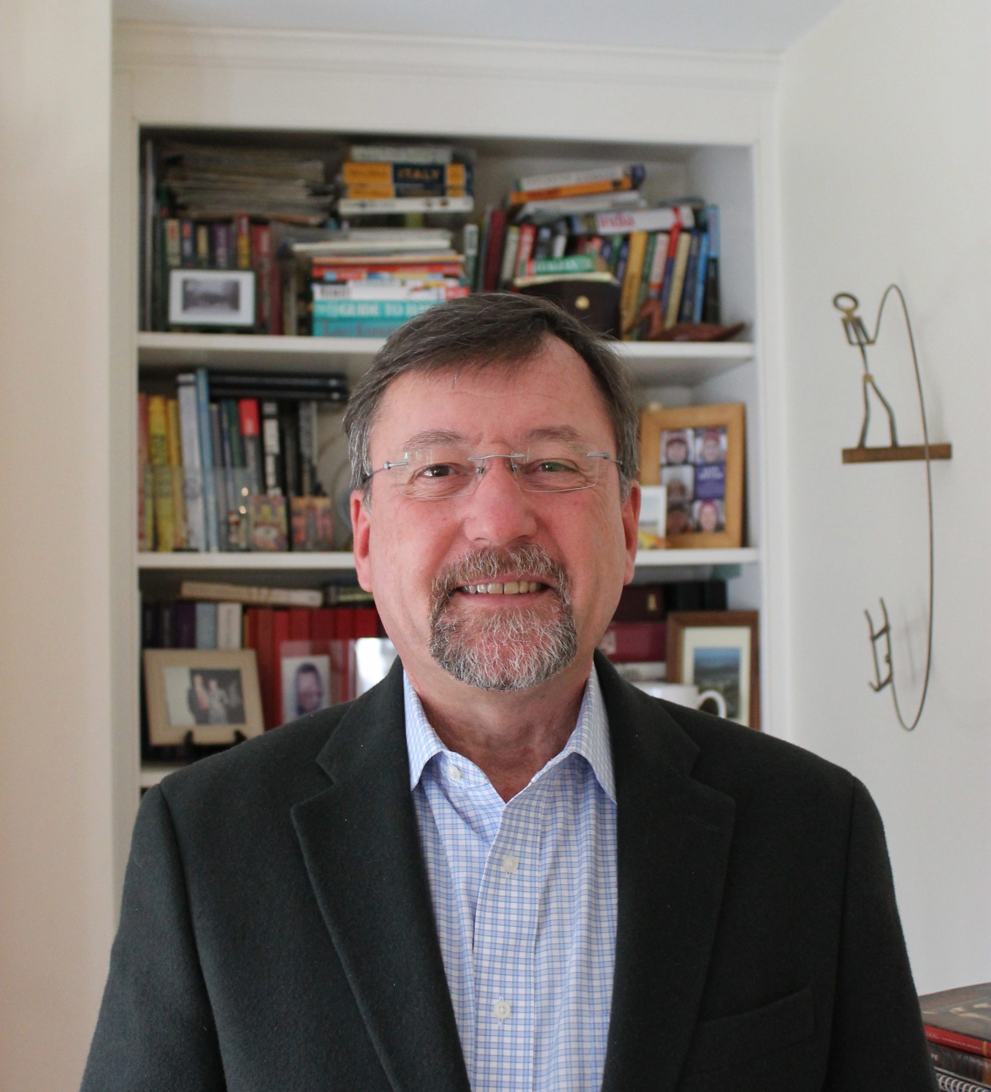 Headshot photo man with wire rim glasses and beard wearing a black coat jacket over a blue and white checked shirt.