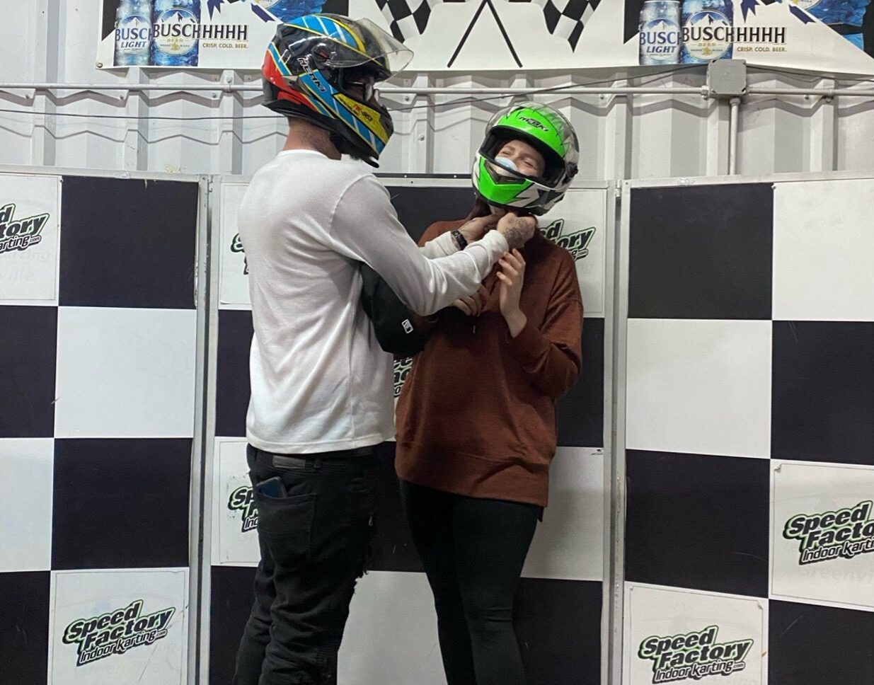 A man and woman in motorcycle helmets stand in front of a black and white checker background.