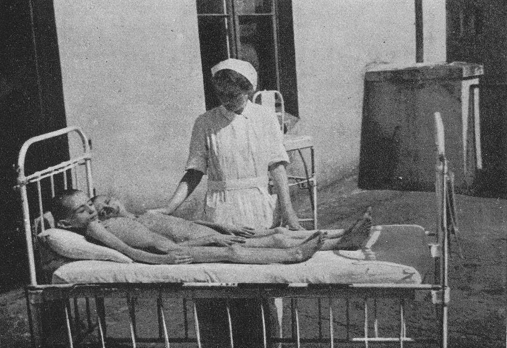 old black and white photo of two very thin children on a bed with a nurse in a white uniform at the bedside. 