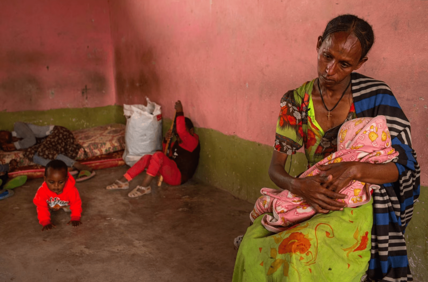 Thin dark skinned woman holds a sick infant while small children play near by