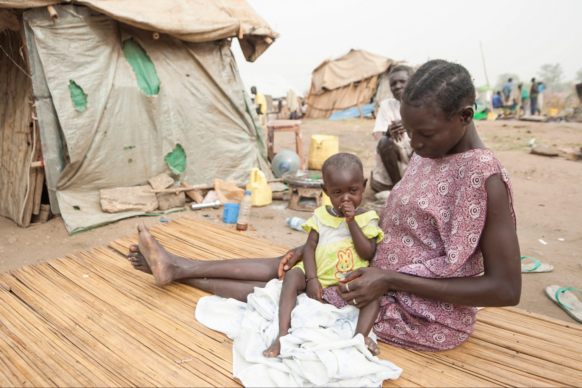 a woman and young child sit on a straw mat outside a canvas tent